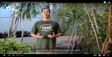 Print de uma cena de um vídeo no You Tube da UFES, mostrando um repórter falando e gesticulando em frente ao jardim de um prédio, com a legenda "O Laboratório de Paleontologia da UFES" e o título do vídeo "Conheça o Laboratório de Paleontologia da Ufes"