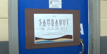 Imagem retirado do vídeo da TV Guarapari mostra a porta azul de uma sala de aula, sobre a qual há um cartaz de papel escrito "Exposição Sambaqui na sala de aula - Culminância do projeto interdisciplinar Tinha Sambaqui aqui: Arqueologia e História Indígena Capixabas"