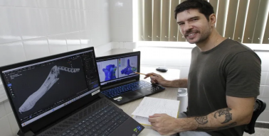 Foto do Richard no laboratório, em frente a dois computadores. Em um deles, a tela mostra a imagem do crânio digitalizado do Anhanguera piscator. No outro, mostra duas vértebras do mesmo animal com cores artificiais, majoritariamente em azul.